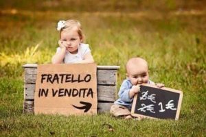 Una bambina piccola sul prato col fratellino e un ironico cartello con la scritta "fratello in vendita"