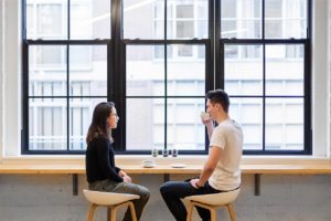 Un ragazzo e una ragazza prendono un caffé assieme e dialogano tra loro e sullo sfondo grandi finestre a riquadri