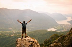 Un giovane uomo sopra una vetta ammira il panorama e alza le braccia in segno di vittoria per aver raggiunto l'obiettivo