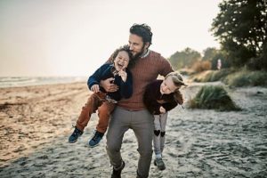 Un giovane papà porta sottobraccio con energia i suoi bambini divertiti in spiaggia