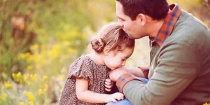 Un papà bacia sulla fronte la sua bambina mentre lei gli dà un bacio sulla mano e sullo sfondo un prato verde e fiorito