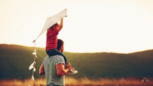 Un papà tiene sulle spalle il suo bambino che regge un aquilone e insieme guardano il cielo verso l'orizzonte