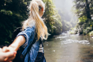 Una ragazza bionda di spalle si avventura nel bosco tenendo per mano qualcuno che vorrebbe trattenerla indietro