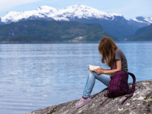 Una ragazza intenta a leggere un libro assegnato come compito per le vacanze e sullo sfondo una montagna innevata che si specchia in un lago blu