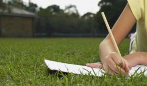 La mano di un ragazzo che fa i compiti delle vacanze con matita e carta appoggiata su un prato verde