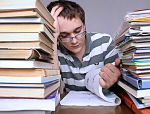Un ragazzo con gli occhiali e una mano tra i capelli intento a studiare tra due pile di libri