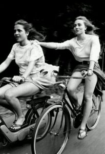 Una ragazza in motorino ne traina un'altra in bicicletta gambe al vento nell'ultimo giorno di scuola degli anni 80