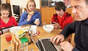 Una famiglia papà mamma figlio figlia mangiano sul tavolo con cellulare tablet smartphone cuffie