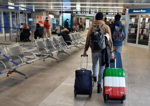 Un giovane all'aeroporto di spalle porta con sé due trolley di cui uno con i colori della bandiera italiana