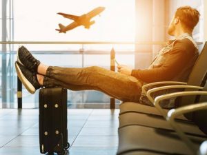 Un giovane attende il suo volo all'aeroporto con i piedi sulla sua valigia e sullo sfondo un aereo in fase di decollo
