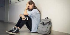 Una ragazza seduta a terra sul pavimento della scuola con lo zaino di fianco e il cellulare in mano in preda al panico da studio